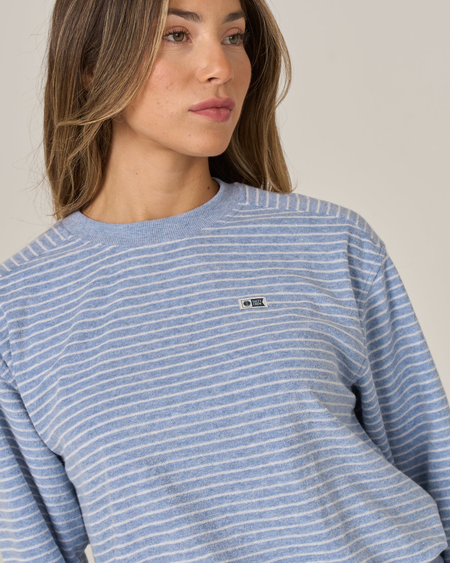 A woman with light brown hair is seen looking slightly to the side against a plain background, wearing the Salty Crew Leeward Crew in Blue Heather—a blue and white striped crew neck pullover crafted from a soft cotton-polyester blend and featuring a small Salty Crew branded patch on the chest.