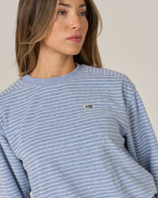 A woman with light brown hair is seen looking slightly to the side against a plain background, wearing the Salty Crew Leeward Crew in Blue Heather—a blue and white striped crew neck pullover crafted from a soft cotton-polyester blend and featuring a small Salty Crew branded patch on the chest.