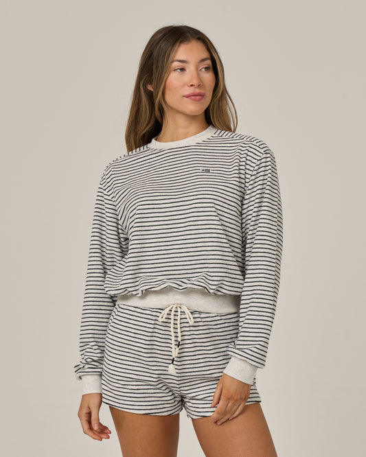 A woman models the Salty Crew Leeward Crew - Light Heather, a matching striped long-sleeve crew neck sweatshirt and drawstring shorts set crafted from a soft cotton-polyester blend. She stands against a plain light background, exuding a relaxed yet stylish vibe as she gazes slightly to the side.