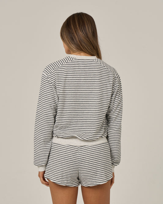 A woman stands with her back to the camera, wearing Salty Crew’s Leeward Crew in Light Heather—a long-sleeved crew neck top and matching shorts. Her brown hair falls loose as she poses against a plain beige background. This set, crafted from a soft cotton-polyester blend, combines comfort with versatile style.