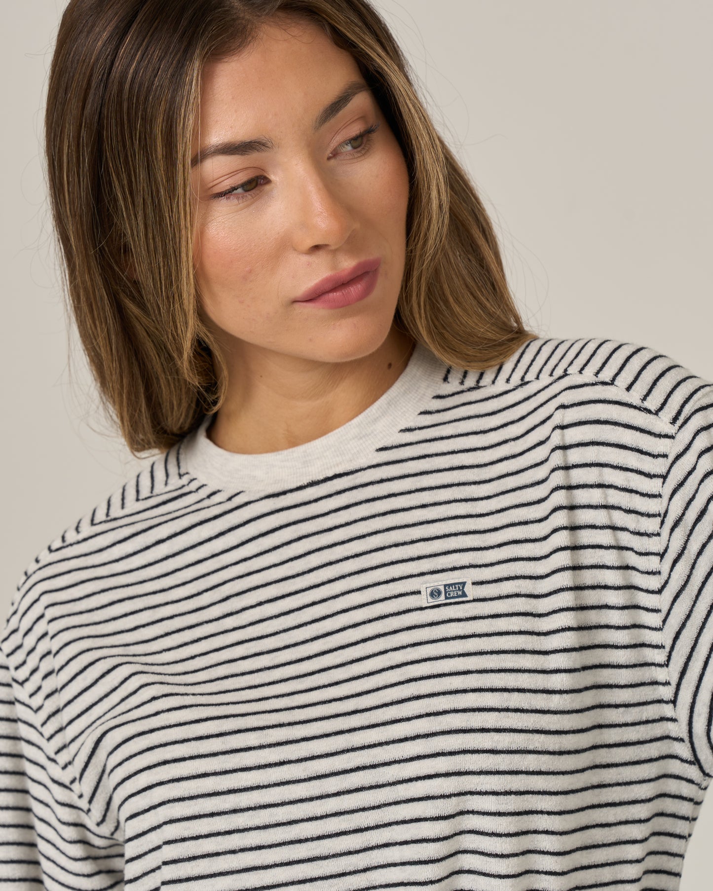 A woman with long brown hair wearing the Salty Crew Leeward Crew in Light Heather, featuring a classic crew neck and crafted from a soft cotton-polyester blend, looks to her left. The sweater stands out against the plain, light-colored background.