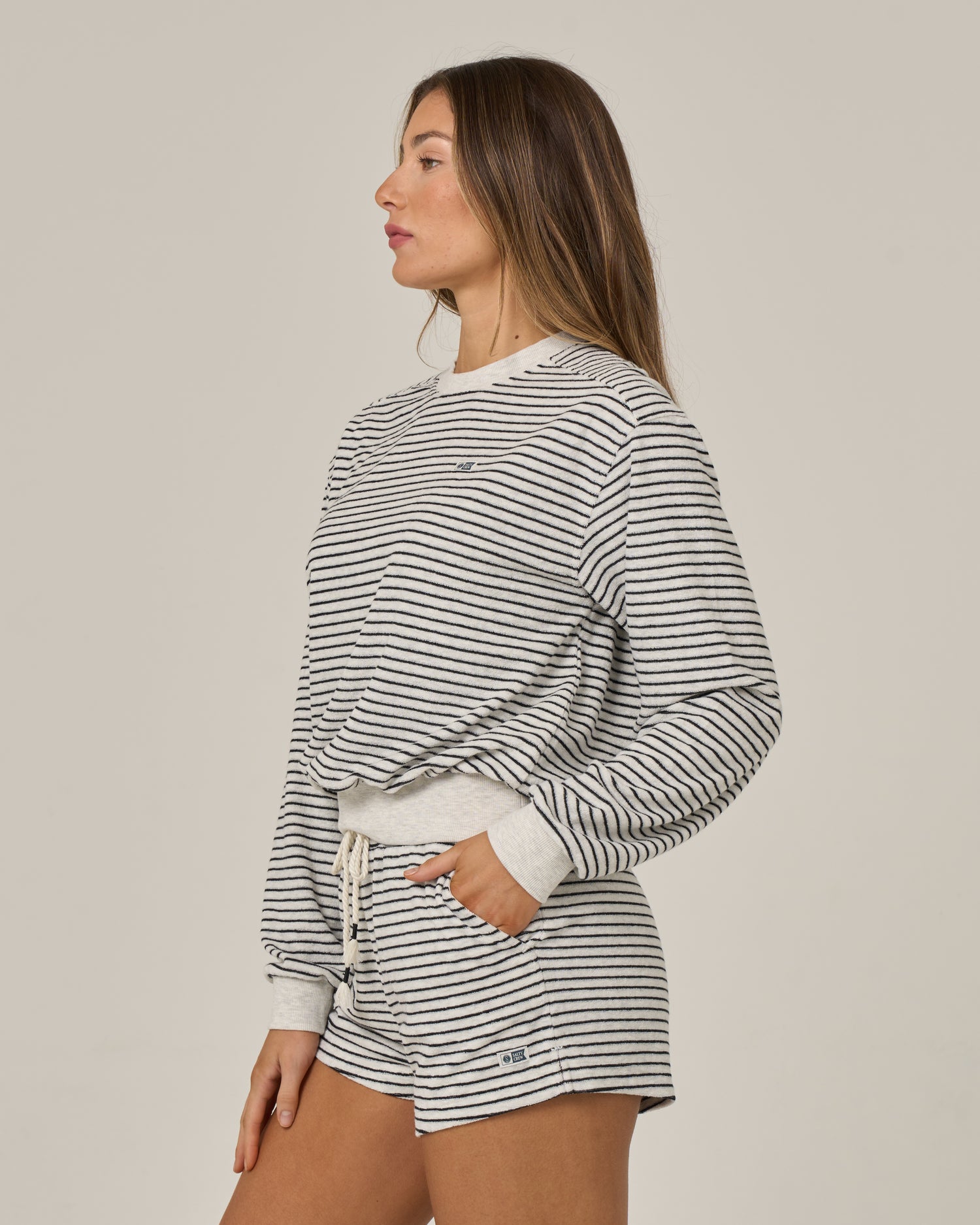 A woman in side profile wears the Salty Crew Leeward Crew - Light Heather, a comfortable cotton-polyester long-sleeve pullover with a crew neck, paired with matching drawstring-waist shorts, both featuring stylish black-and-white stripes. She stands against a plain light background.