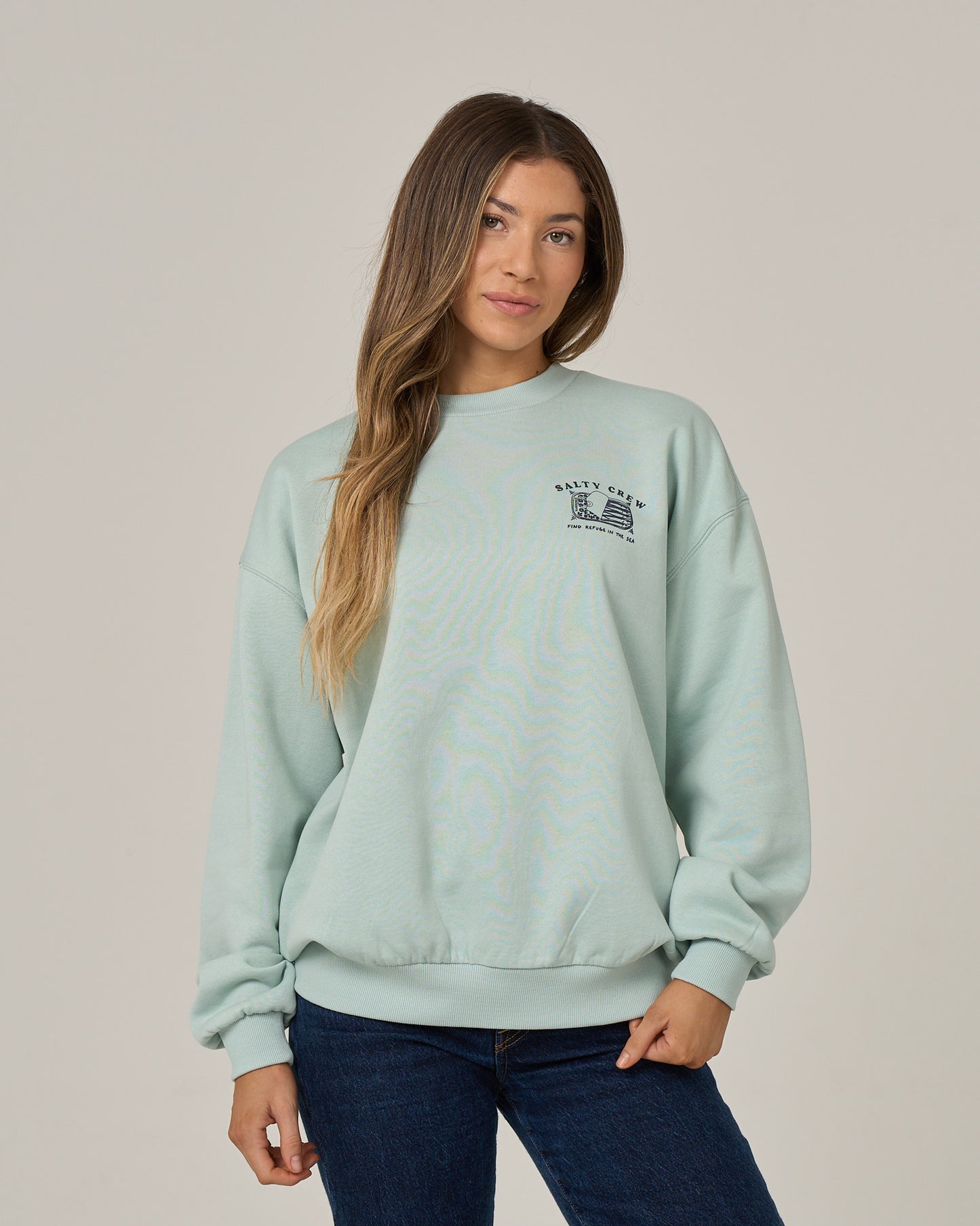 A woman with long hair wears the Salty Crew Cute Tin Crew in Cloud Blue, an oversized cotton-polyester fleece pullover, paired with dark jeans. She stands facing forward with one hand in her pocket against a plain light background.