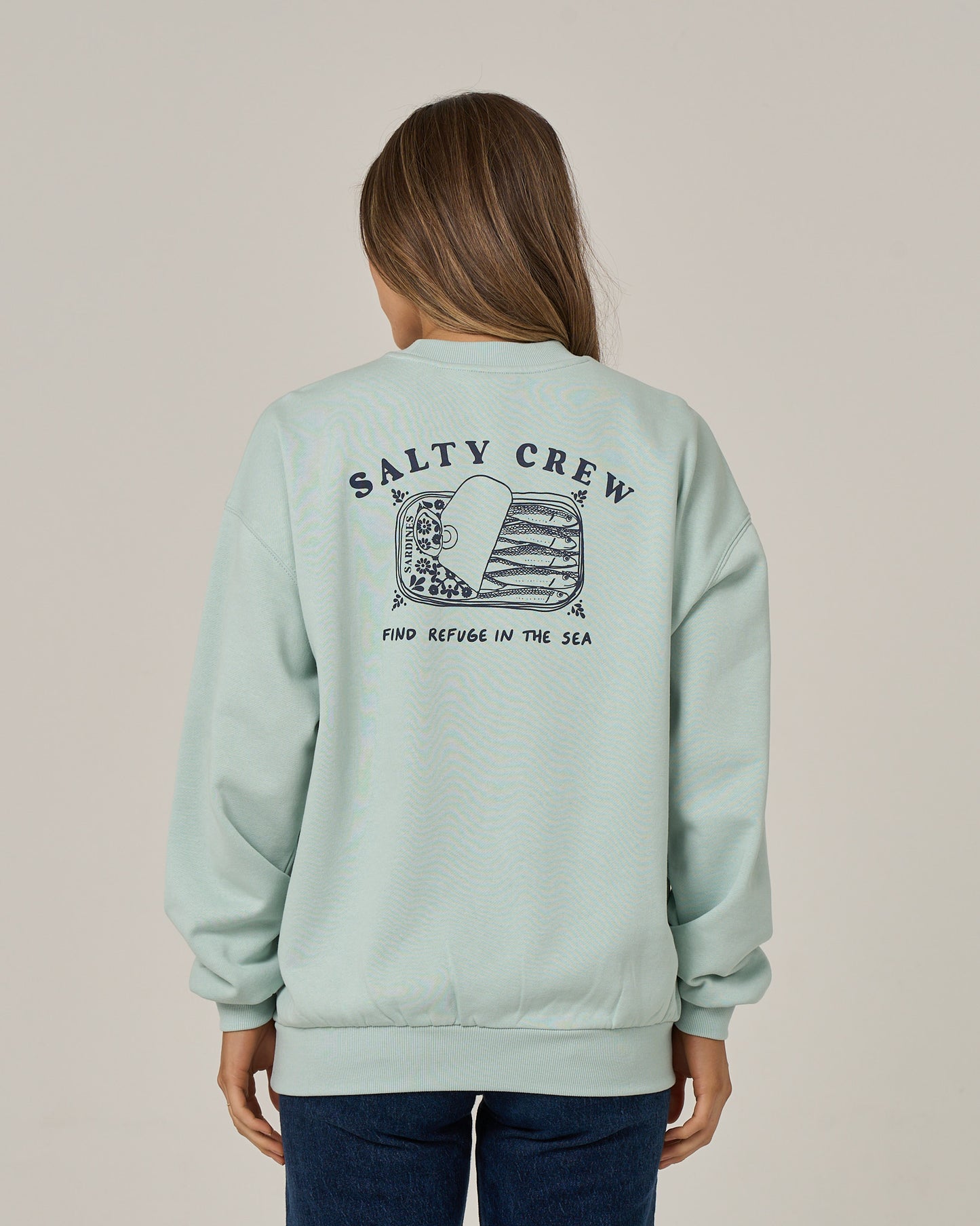A woman with long brown hair, facing away from the camera, wears the Salty Crew Cute Tin Crew in Cloud Blue—an oversized crew neck pullover crafted from soft cotton polyester fleece. The sweatshirt features a sea-themed graphic and “FIND REFUGE IN THE SEA” screen printed on the back.