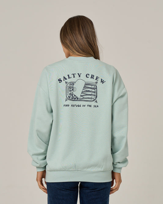 A woman with long brown hair, facing away from the camera, wears the Salty Crew Cute Tin Crew in Cloud Blue—an oversized crew neck pullover crafted from soft cotton polyester fleece. The sweatshirt features a sea-themed graphic and “FIND REFUGE IN THE SEA” screen printed on the back.