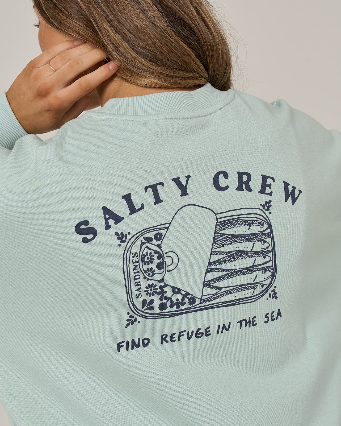 A person wearing the Salty Crew Cute Tin Crew in Cloud Blue, an oversize crew neck pullover made from soft cotton-polyester fleece. The sweatshirt features a sardine can graphic and the words “Salty Crew” and “Find Refuge in the Sea” screen printed on the back, combining cozy comfort with bold style.