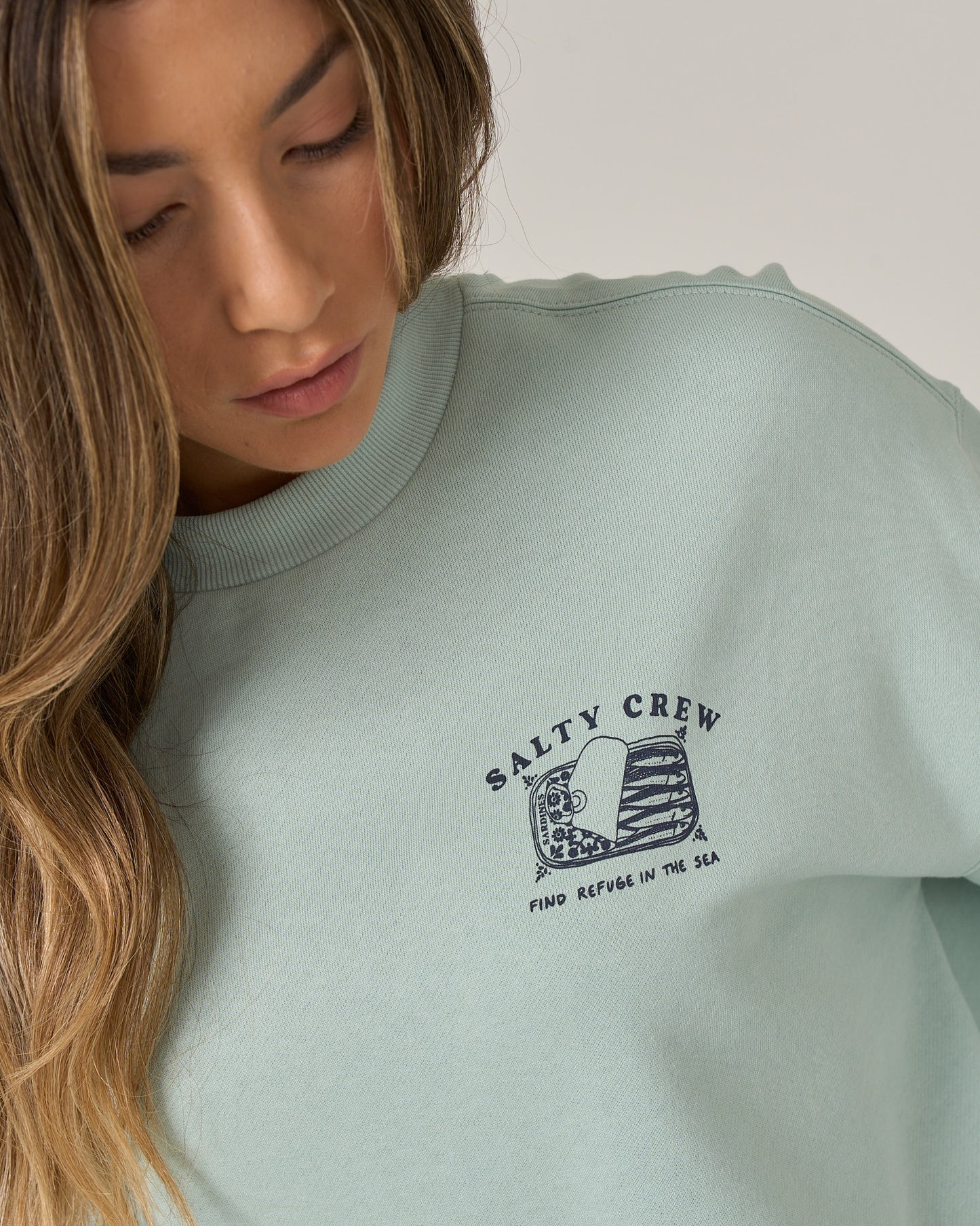 A woman with long, wavy hair looks down while wearing a cloud blue Salty Crew Cute Tin Crew oversize crew neck pullover, featuring a small screen print graphic and the words “Find Refuge in the Sea” on the chest, crafted from soft cotton polyester fleece.