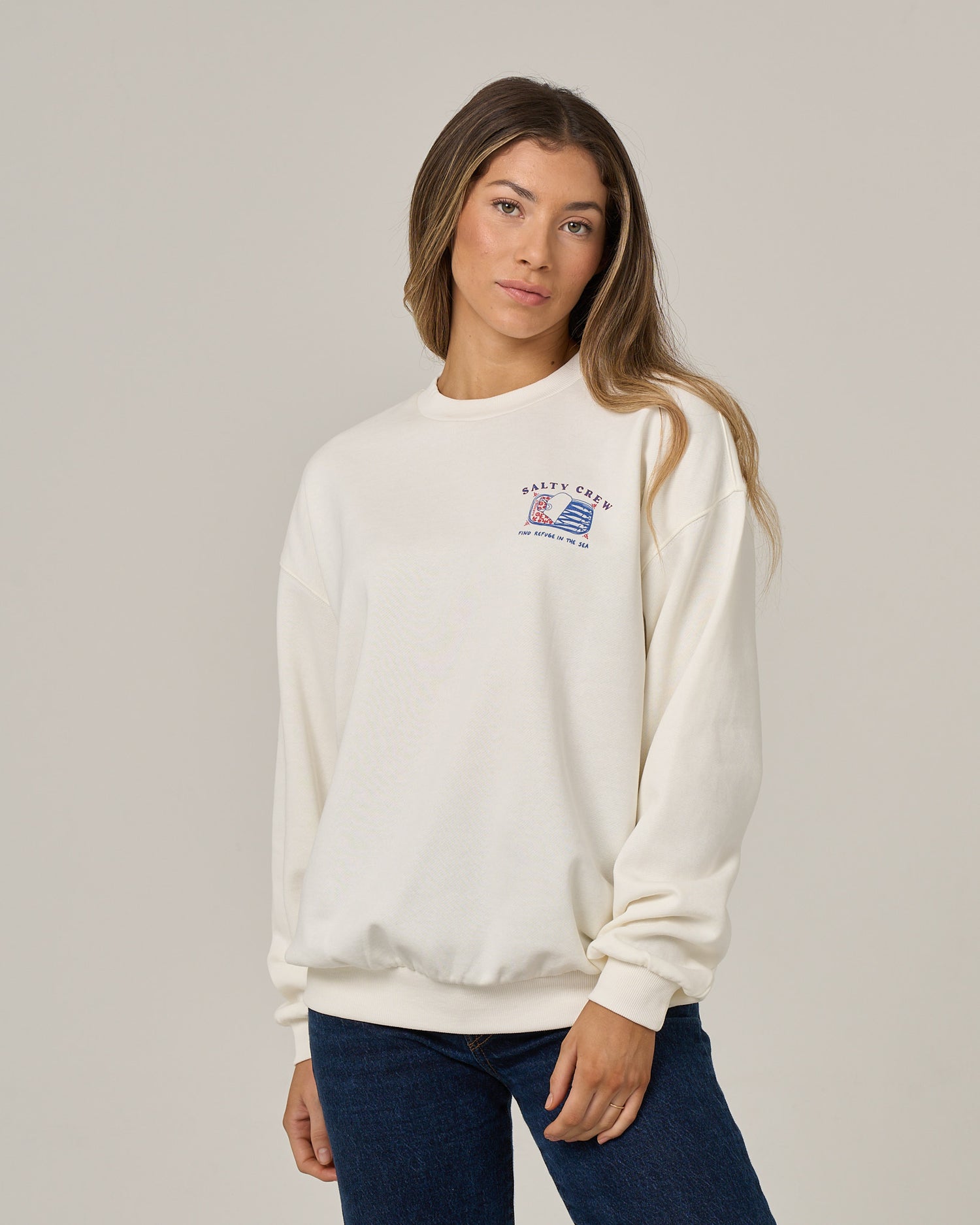 A woman with long hair wears the Salty Crew Cute Tin Crew in off white—an oversized crew neck pullover crafted from soft cotton polyester fleece, distinguished by a small screen-printed graphic and text on the left chest. She pairs it with blue jeans and stands against a plain light grey background.