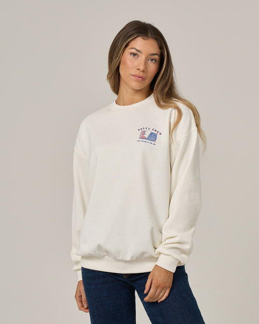 A woman with long hair wears the Salty Crew Cute Tin Crew in off white—an oversized crew neck pullover crafted from soft cotton polyester fleece, distinguished by a small screen-printed graphic and text on the left chest. She pairs it with blue jeans and stands against a plain light grey background.