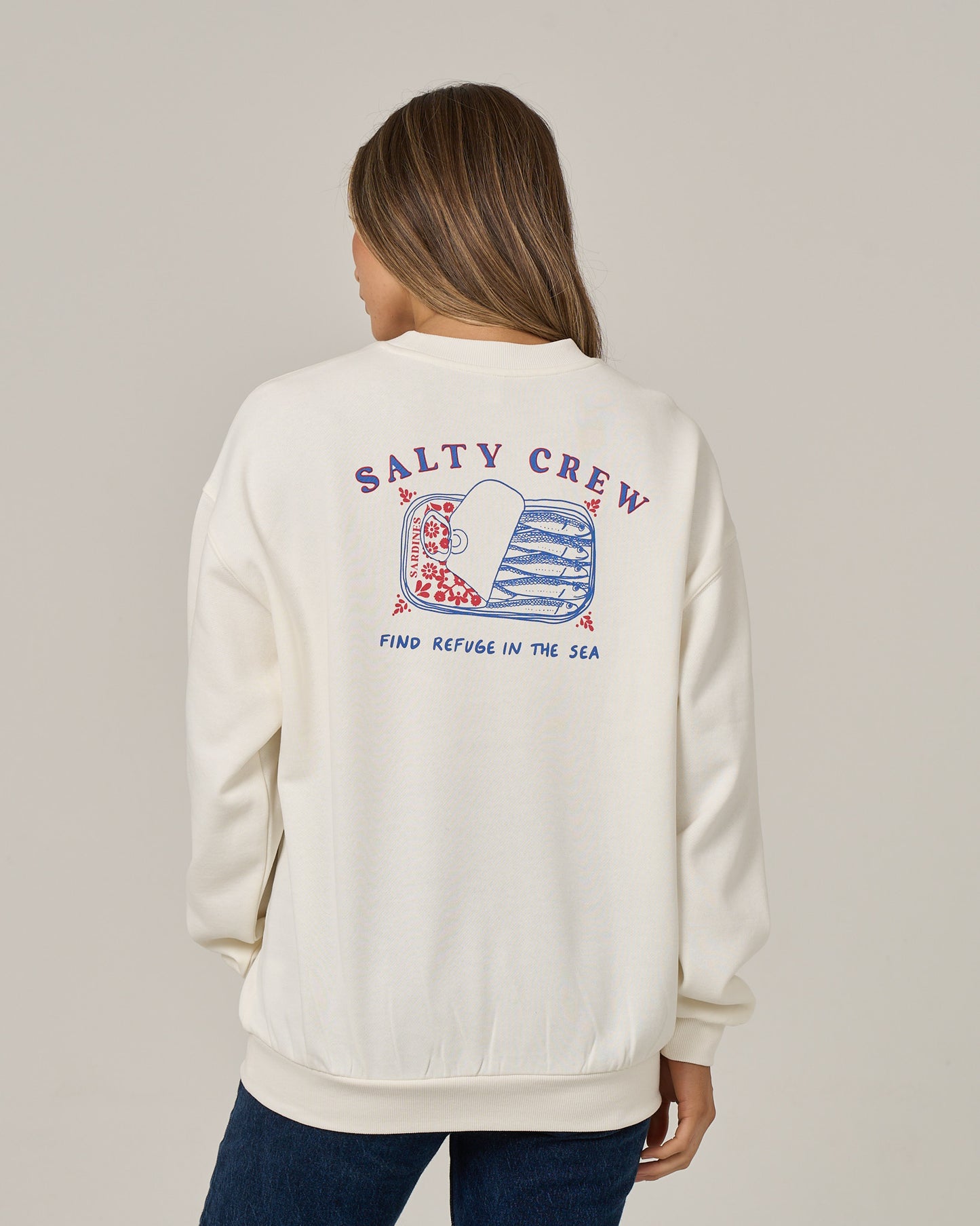 A woman facing away, wearing the Salty Crew Cute Tin Crew in Off White—an oversized crew neck sweatshirt made from soft cotton-polyester fleece, featuring “SALTY CREW” and “FIND REFUGE IN THE SEA” printed on the back in red and blue alongside a graphic of a fish and a boat.