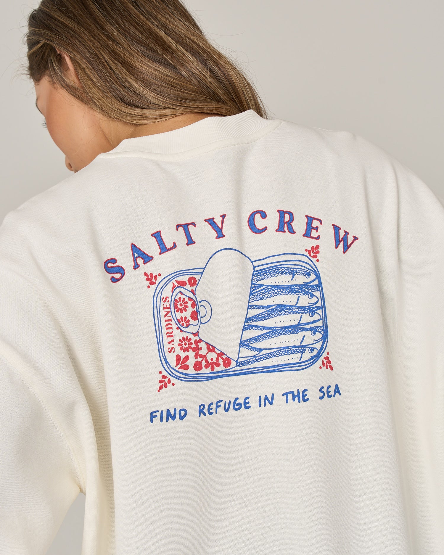 A person wearing the Salty Crew Cute Tin Crew in Off White, an oversized crew neck pullover sweatshirt featuring a sardine tin screen print graphic, SALTY CREW text, and “FIND REFUGE IN THE SEA” written below on the back, crafted from a soft cotton-polyester fleece blend.