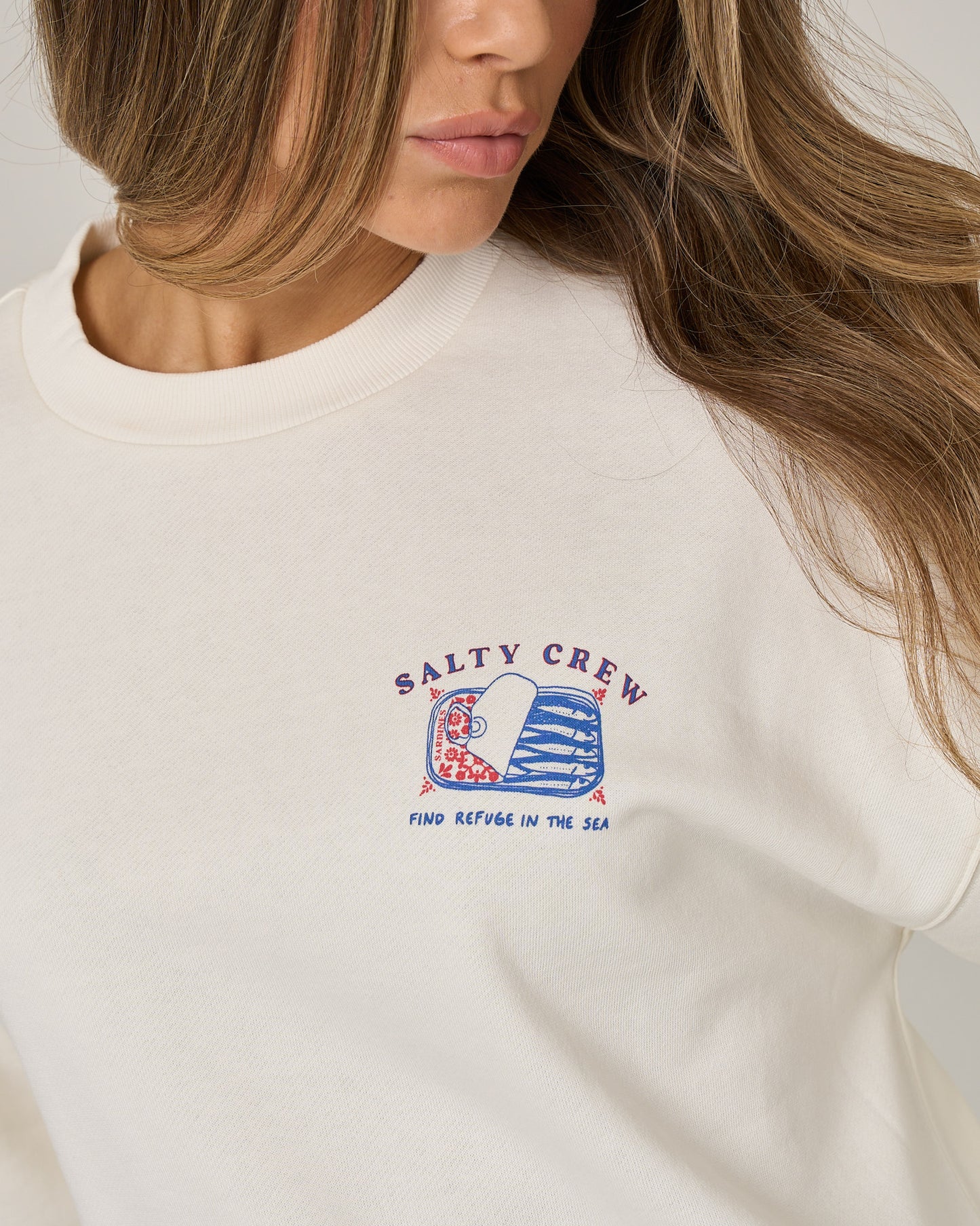 Wearing the Salty Crew Cute Tin Crew - Off White, a woman sports an oversize crew neck pullover featuring a wave graphic and “Find Refuge in the Sea” screen printed on the chest. Crafted from soft cotton-polyester fleece, this white sweatshirt by Salty Crew complements her loose hair as her face stays partially visible.