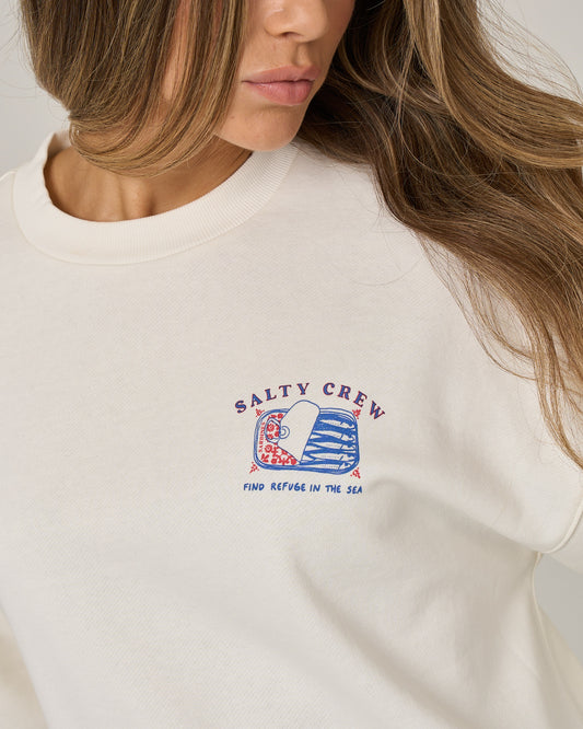 Wearing the Salty Crew Cute Tin Crew - Off White, a woman sports an oversize crew neck pullover featuring a wave graphic and “Find Refuge in the Sea” screen printed on the chest. Crafted from soft cotton-polyester fleece, this white sweatshirt by Salty Crew complements her loose hair as her face stays partially visible.