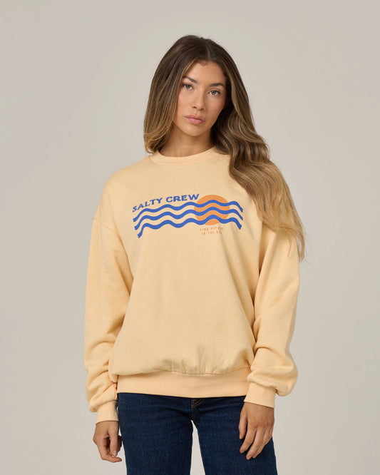 A woman with long brown hair poses in the Salty Crew Boat Legs Crew – Custard, an oversized crew neck pullover sweatshirt made from soft cotton-polyester fleece. The top features a bold screen print of a blue wave and orange sun graphic with the words “Salty Crew, Find Refuge In The Sea.” She pairs it with blue jeans while standing against a plain light gray background.