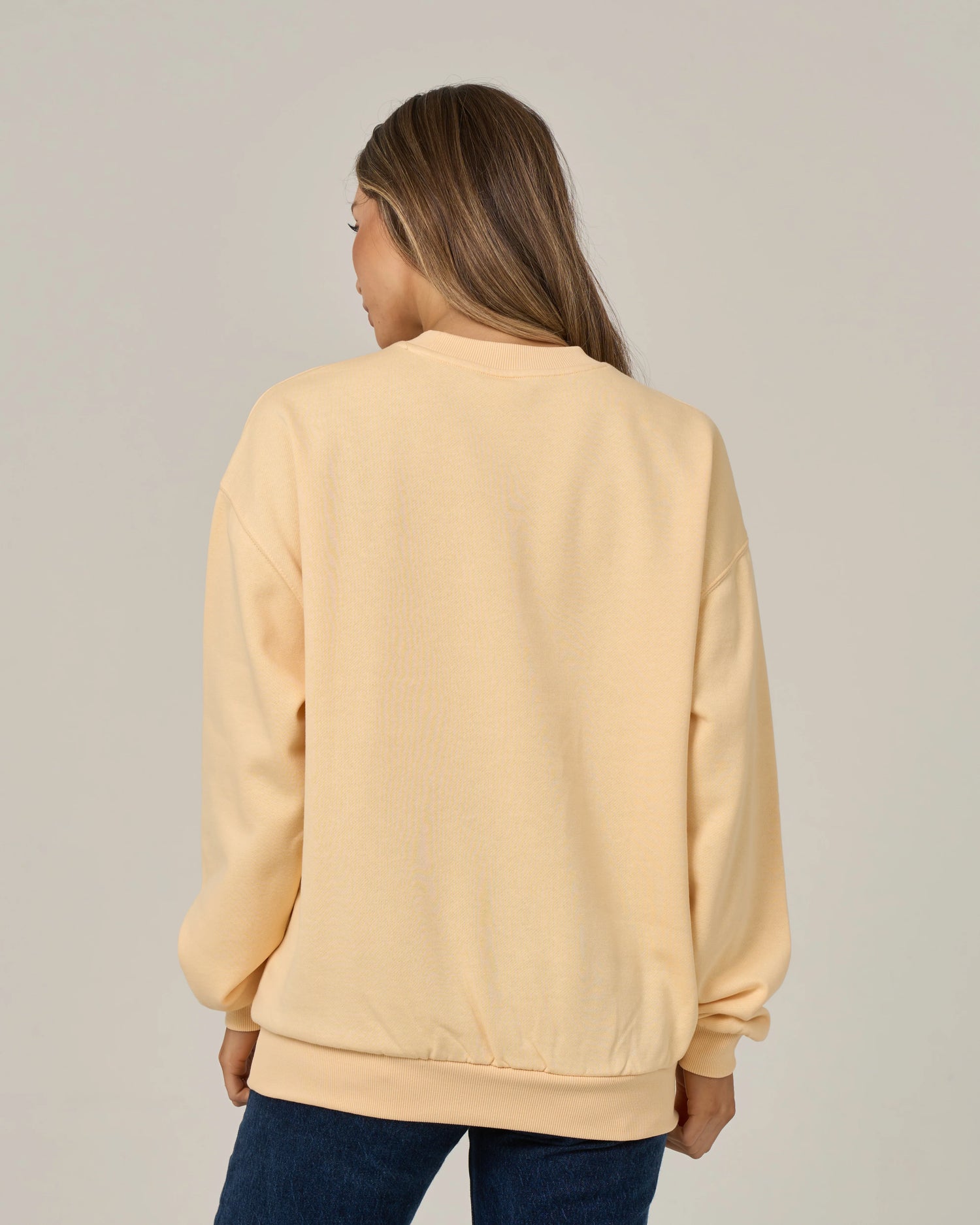 A person with long brown hair is standing with their back to the camera, wearing the Salty Crew Boat Legs Crew in Custard—a light beige oversized cotton-polyester fleece pullover—and dark blue jeans against a plain light background.