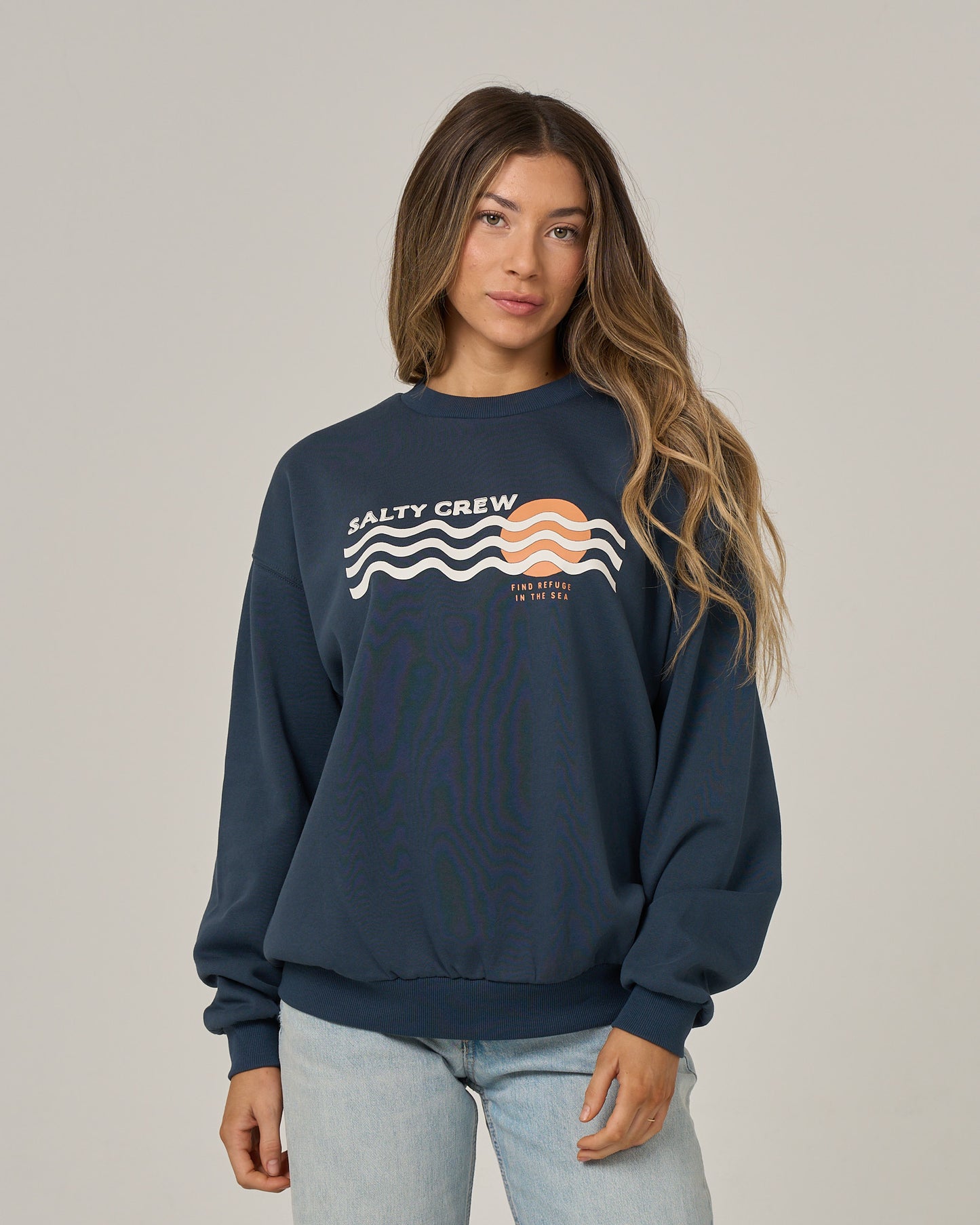 A woman with long brown hair wears the Salty Crew Boat Legs Crew in Navy, an oversized crew neck pullover crafted from soft cotton fleece. The sweatshirt features a bold screen print of an orange sun rising over white waves alongside the text “FIND REFUGE IN THE SEA.” She pairs it with light-wash jeans and stands against a plain, light gray studio background.
