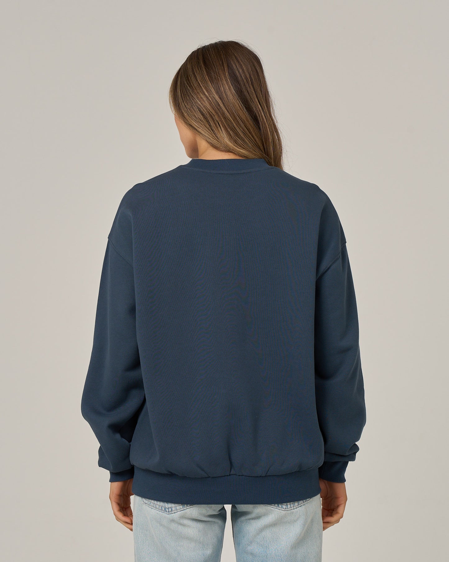 A person with long brown hair, wearing the Salty Crew “Boat Legs Crew - Navy” oversize pullover and light blue jeans, is standing with their back to the camera against a plain light background.