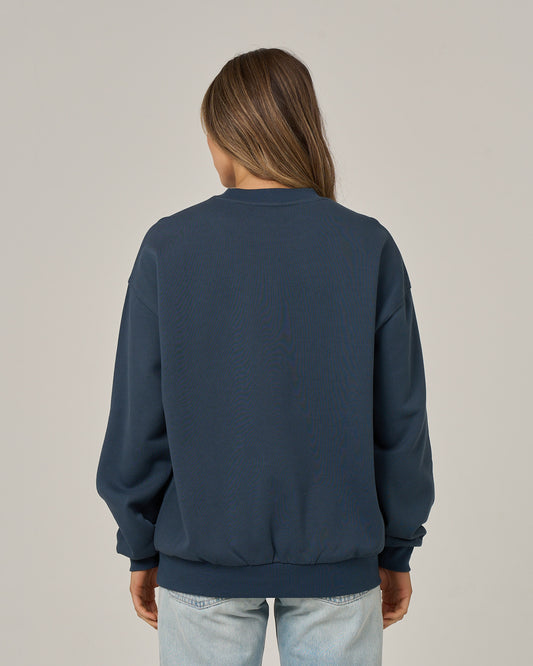A person with long brown hair, wearing the Salty Crew “Boat Legs Crew - Navy” oversize pullover and light blue jeans, is standing with their back to the camera against a plain light background.