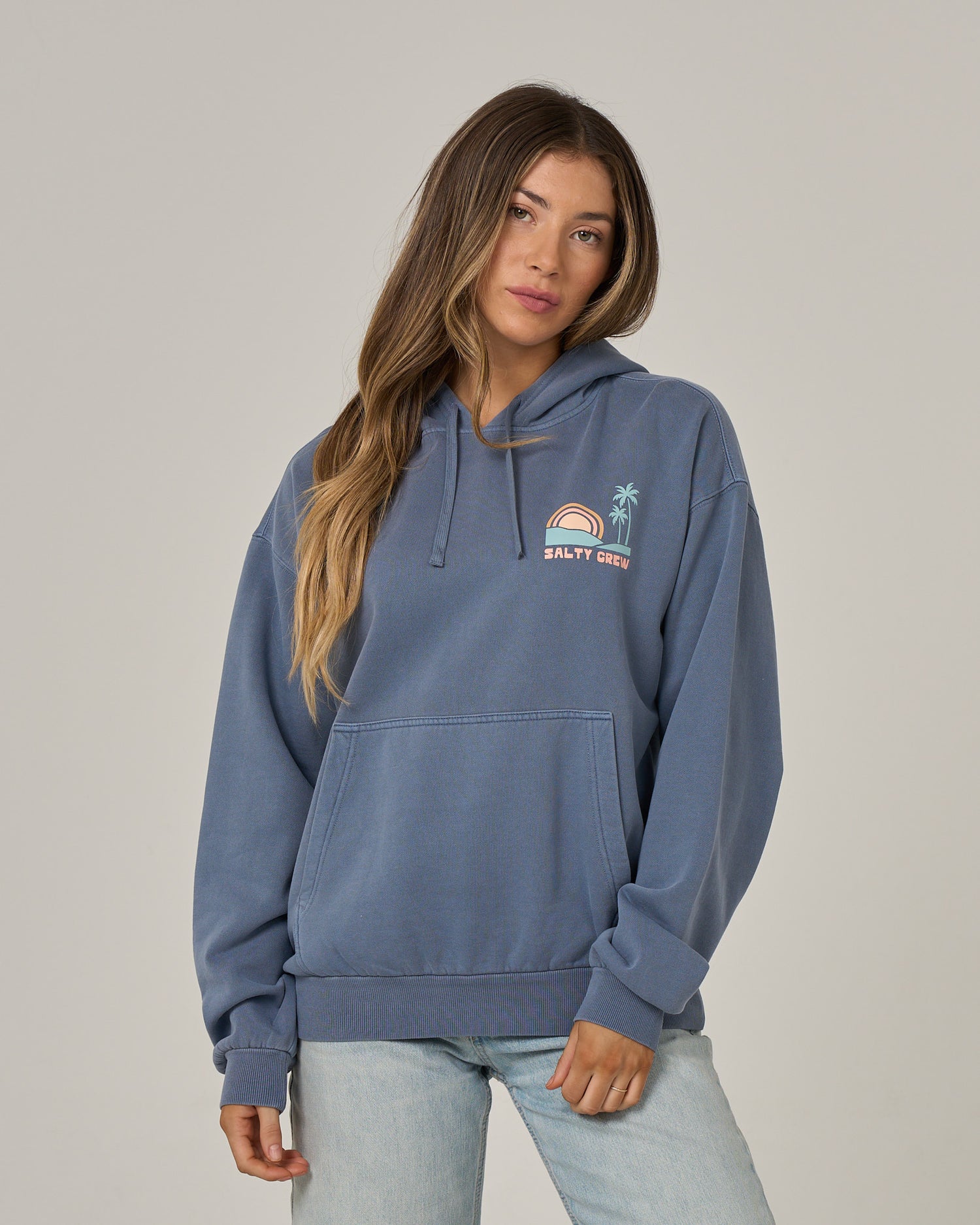 A person with long hair wears the Salty Crew Sushi Roll Hoodie in Flint—an oversized blue fleece hoodie featuring a small rainbow, sun, and palm tree graphic on the chest and a classic kangaroo pocket—paired with light blue jeans, standing against a plain light background.