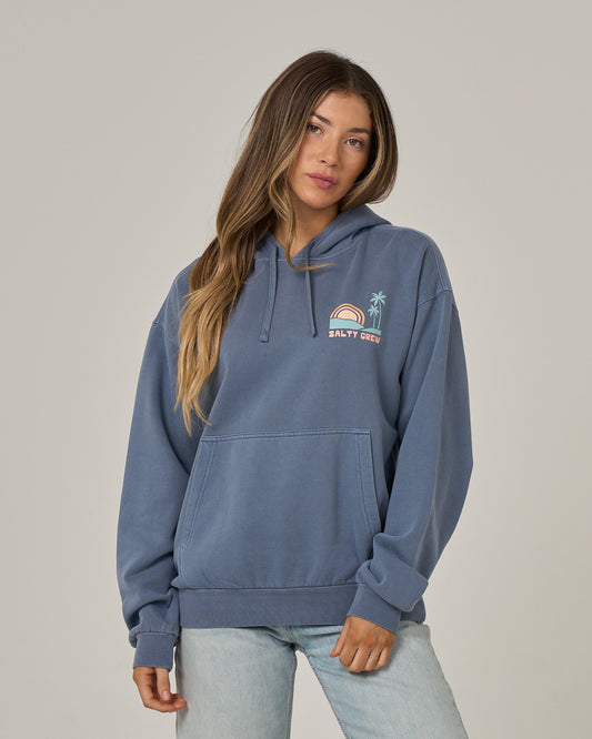 A person with long hair wears the Salty Crew Sushi Roll Hoodie in Flint—an oversized blue fleece hoodie featuring a small rainbow, sun, and palm tree graphic on the chest and a classic kangaroo pocket—paired with light blue jeans, standing against a plain light background.