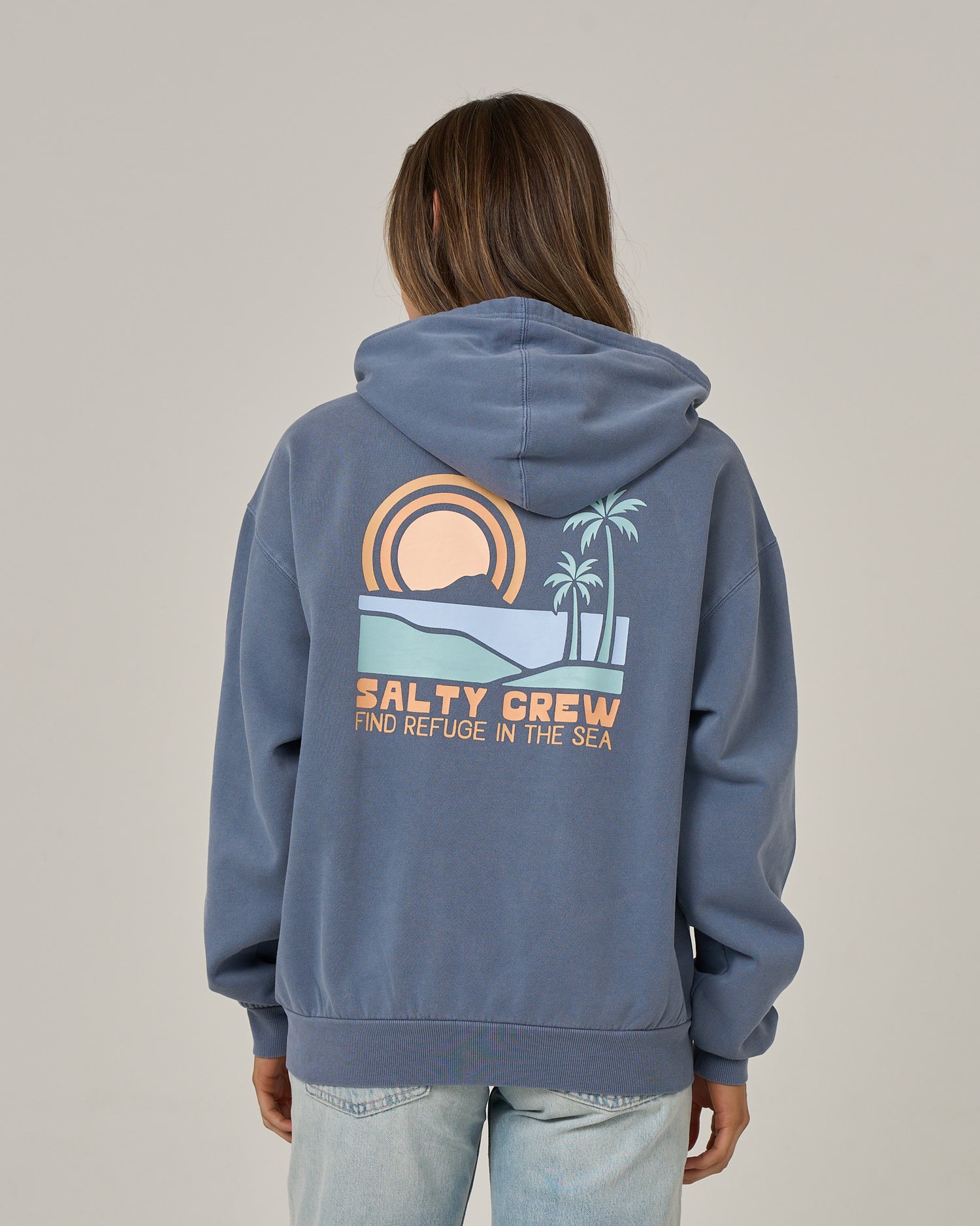 A person wearing the Salty Crew Sushi Roll Hoodie in Flint, featuring a sunset, palm trees, and the text “FIND REFUGE IN THE SEA” printed on the back, with a classic kangaroo pocket, standing against a plain background.