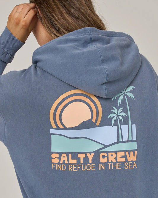 A person wearing the Salty Crew Sushi Roll Hoodie in Flint, an oversized blue fleece hoody featuring a graphic of a sunset, palm trees, and hills with the text “FIND REFUGE IN THE SEA.” The cozy design includes a classic kangaroo pocket. The person is facing away and has one hand touching their head.
