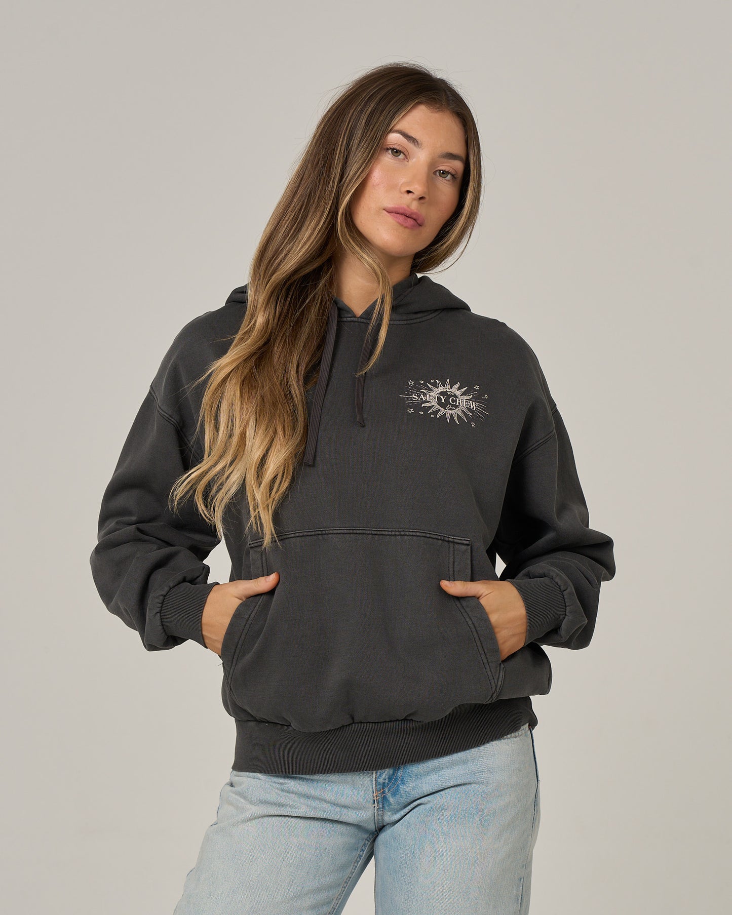 A woman with long, wavy hair stands against a plain light background, hands tucked into the front pocket of her Salty Crew Celestial Sun Hoodie in Faded Black. Featuring a sun and moon graphic on the chest, this oversized hoodie pairs effortlessly with light jeans. The vintage wash and soft fleece ensure it’s as comfortable as it is stylish.