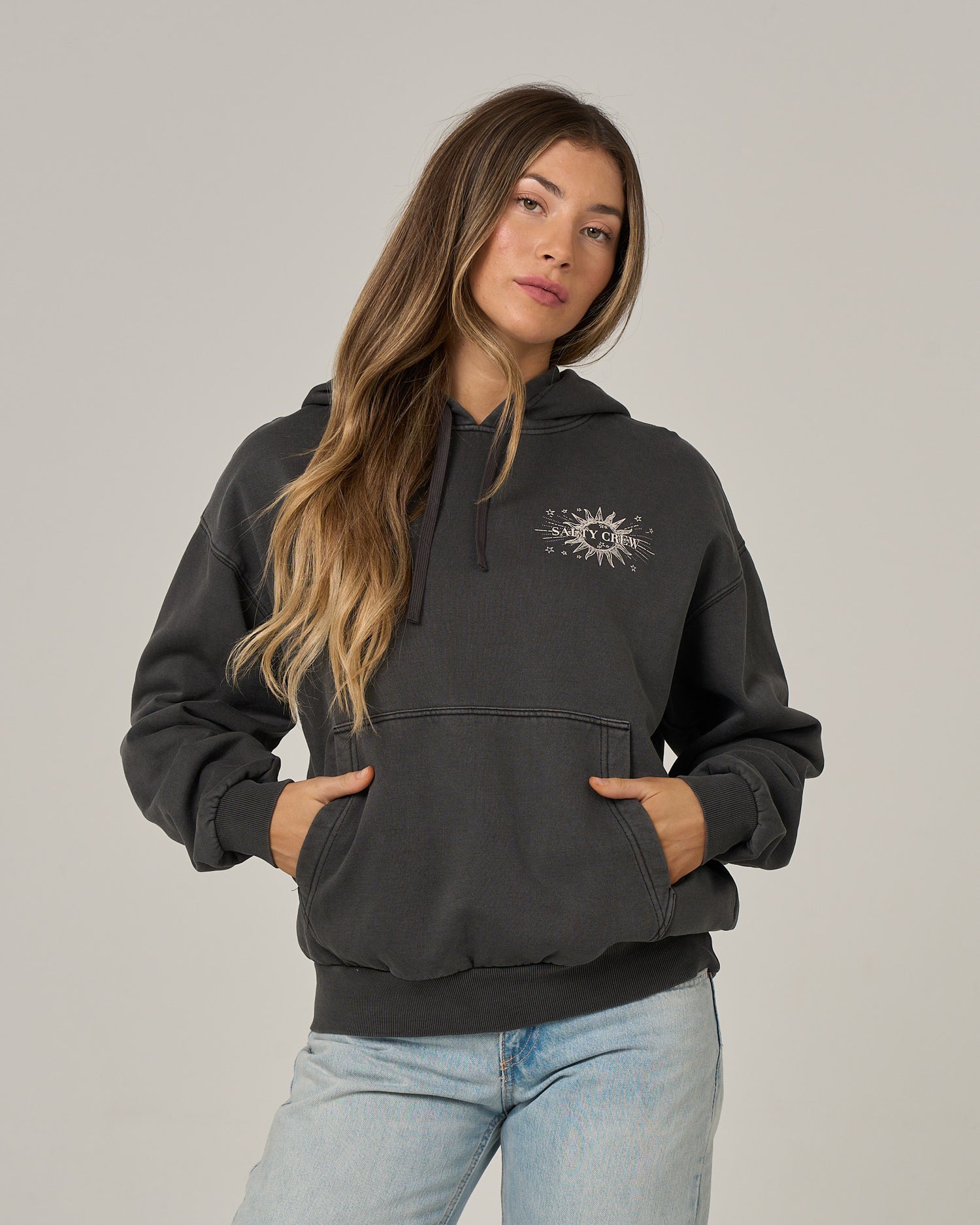 A woman with long, wavy hair stands against a plain light background, hands tucked into the front pocket of her Salty Crew Celestial Sun Hoodie in Faded Black. Featuring a sun and moon graphic on the chest, this oversized hoodie pairs effortlessly with light jeans. The vintage wash and soft fleece ensure it’s as comfortable as it is stylish.