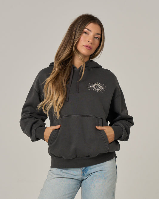 A woman with long, wavy hair stands against a plain light background, hands tucked into the front pocket of her Salty Crew Celestial Sun Hoodie in Faded Black. Featuring a sun and moon graphic on the chest, this oversized hoodie pairs effortlessly with light jeans. The vintage wash and soft fleece ensure it’s as comfortable as it is stylish.