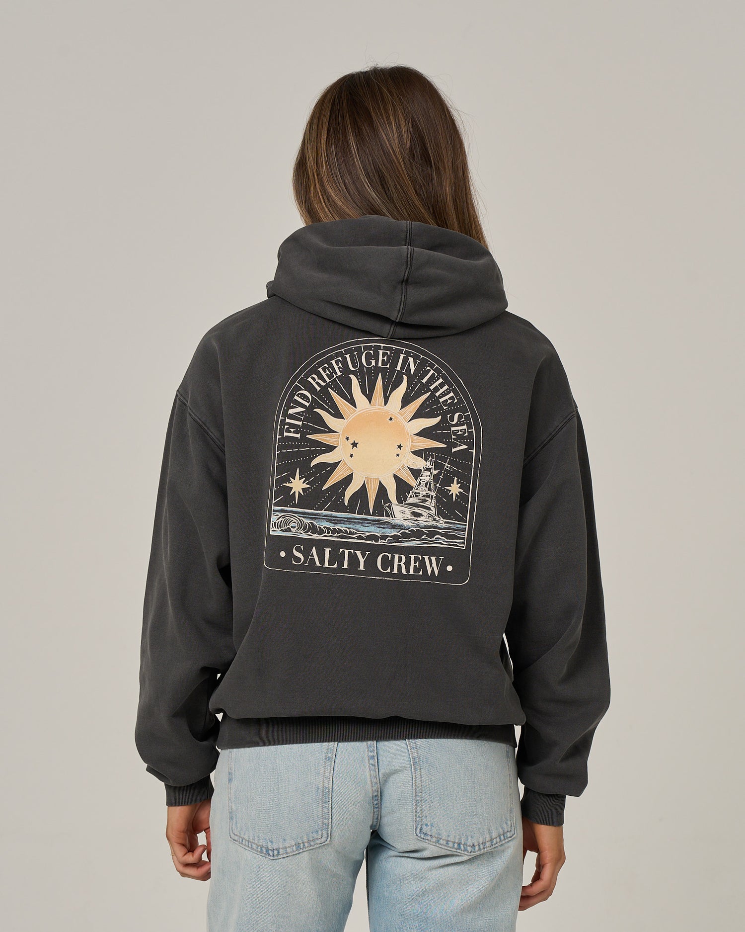 A person wearing the Salty Crew Celestial Sun Hoodie in faded black, featuring a sun and sea graphic on the back with the words “Find Refuge in the Sea” and “Salty Crew,” paired with light blue jeans, stands facing away from the camera.