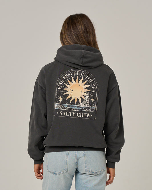 A person wearing the Salty Crew Celestial Sun Hoodie in faded black, featuring a sun and sea graphic on the back with the words “Find Refuge in the Sea” and “Salty Crew,” paired with light blue jeans, stands facing away from the camera.