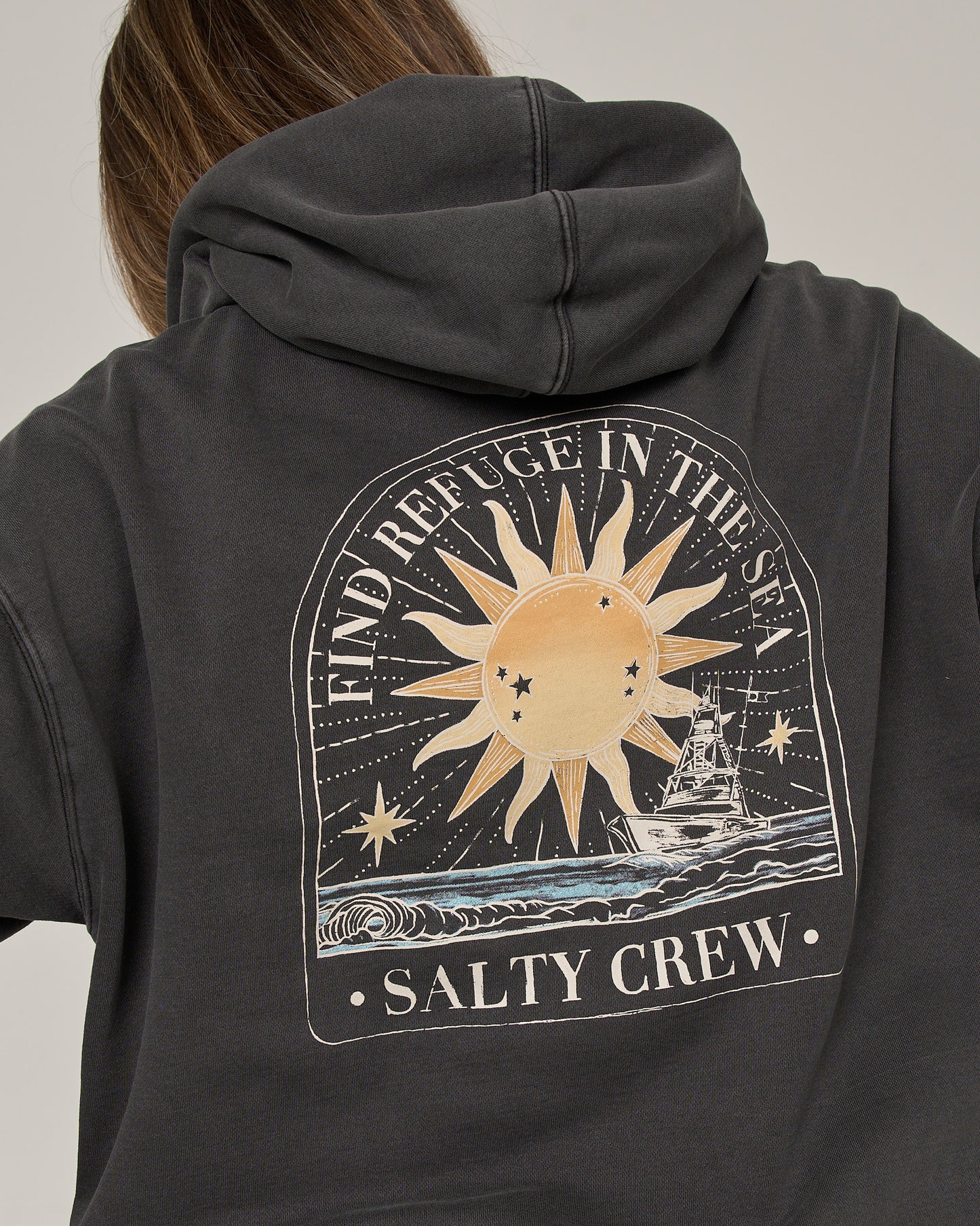 A person wears the Salty Crew Celestial Sun Hoodie in Faded Black, featuring an oversized fit and a sun, stars, ocean waves, and fishing boat graphic. Crafted from comfortable fleece with a vintage wash, it displays the text “FIND REFUGE IN THE SEA” along with “SALTY CREW.”.