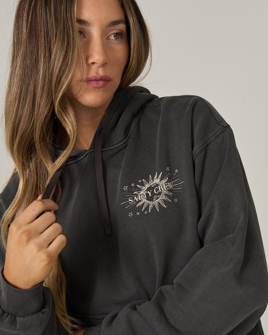 A person with long hair wears the Salty Crew Celestial Sun Hoodie in Faded Black, featuring a sunburst design on the chest. They’re gazing to the side and holding a lock of hair, exuding relaxed style and comfort.