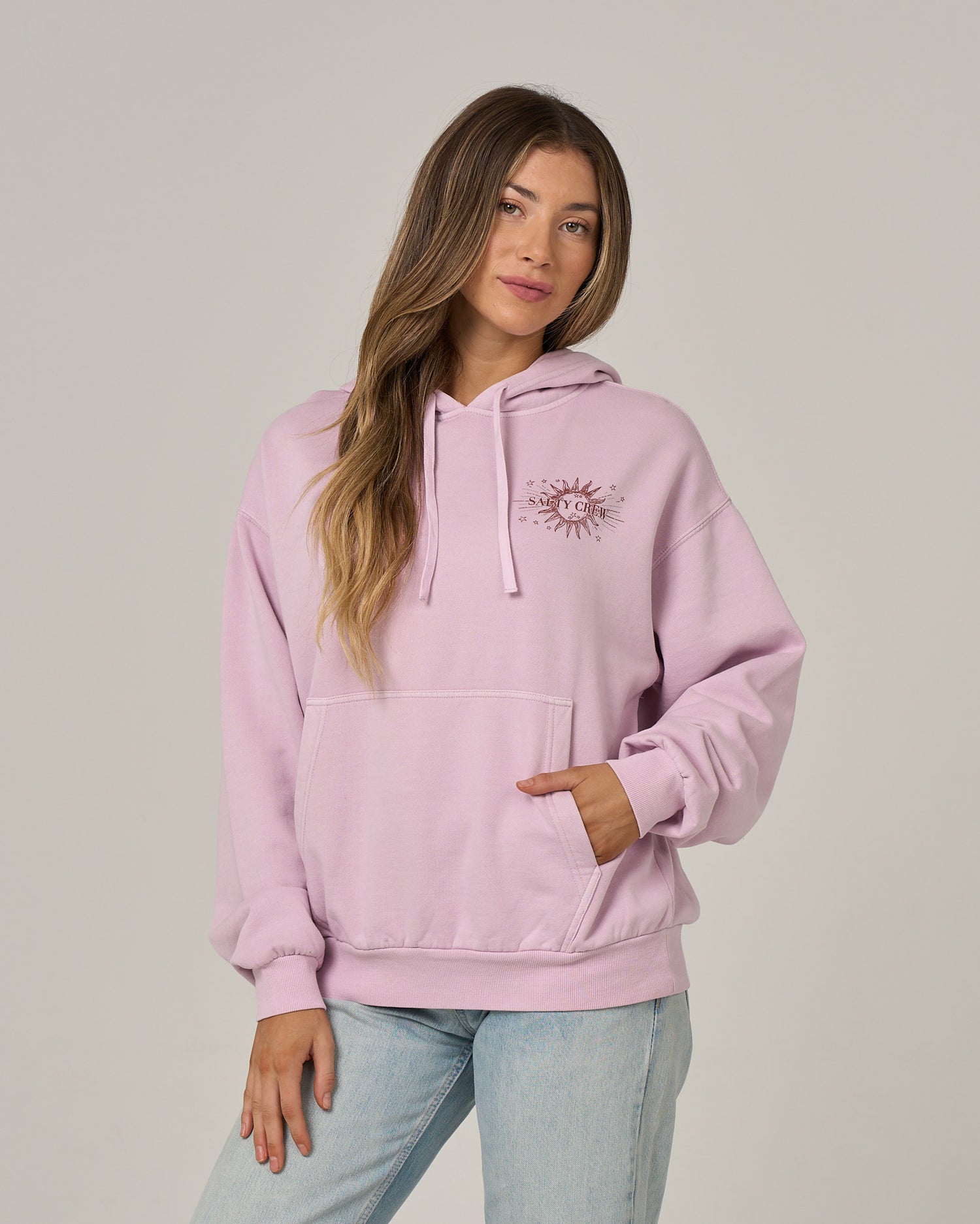 A woman with long, highlighted brown hair poses against a plain background, wearing the Salty Crew Celestial Sun Hoodie in Pale Orchid—a light pink fleece hoodie featuring a sun logo on the chest—paired with light-wash jeans. She has one hand tucked into the front kangaroo pocket.