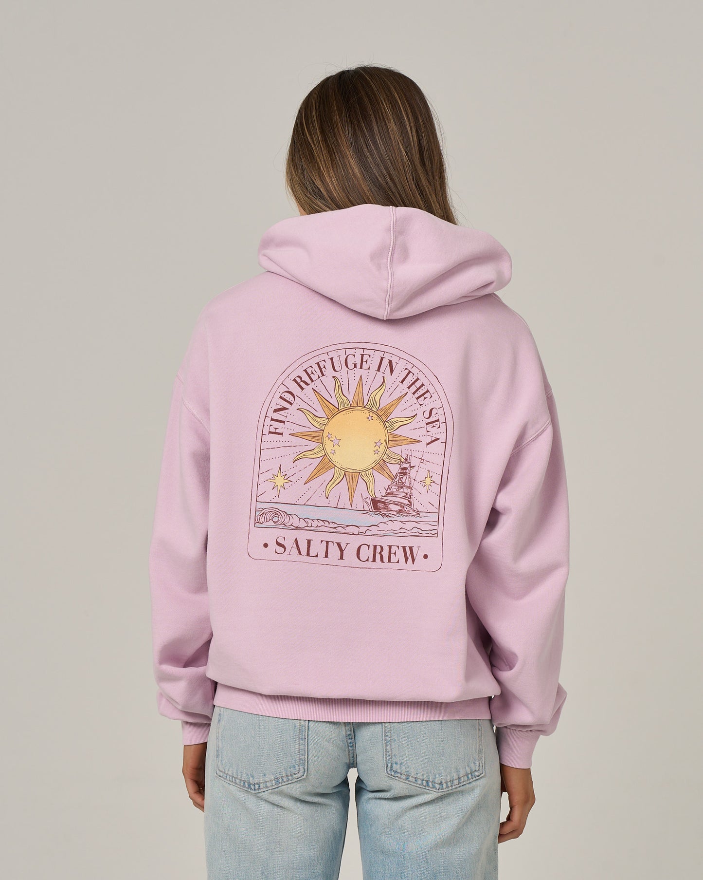 A person stands facing away from the camera, wearing the Salty Crew Celestial Sun Hoodie in Pale Orchid—a light pink, oversize hoodie featuring a sun and ocean graphic on the back with the phrases “FIND REFUGE IN THE SEA” and “SALTY CREW.” The hoodie showcases a vintage wash and cozy fleece construction for ultimate comfort, paired with light blue jeans.