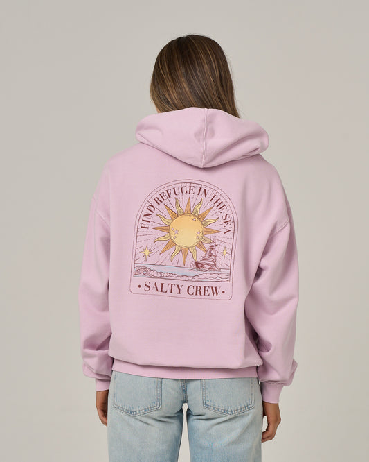 A person stands facing away from the camera, wearing the Salty Crew Celestial Sun Hoodie in Pale Orchid—a light pink, oversize hoodie featuring a sun and ocean graphic on the back with the phrases “FIND REFUGE IN THE SEA” and “SALTY CREW.” The hoodie showcases a vintage wash and cozy fleece construction for ultimate comfort, paired with light blue jeans.