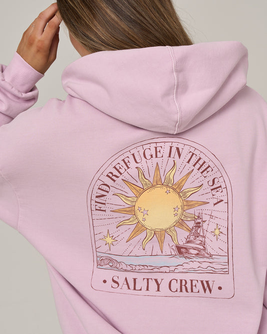 A person wearing the Salty Crew Celestial Sun Hoodie in Pale Orchid, featuring a vintage wash and an oversized fit. The cozy fleece design displays a sun and ocean graphic with stars, a boat on the water, and the words “FIND REFUGE IN THE SEA” alongside “SALTY CREW” on the back.