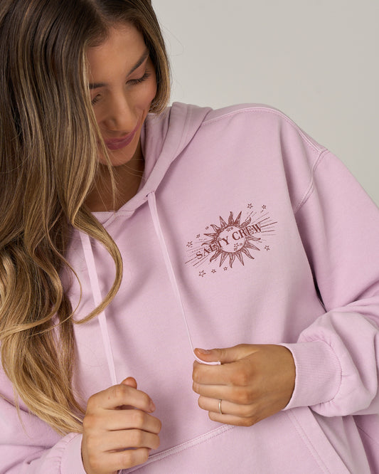 A person with long, wavy hair wearing a light pink Celestial Sun Hoodie in Pale Orchid by Salty Crew, featuring a vintage sun graphic, looks down and smiles gently while holding the soft fleece drawstrings.