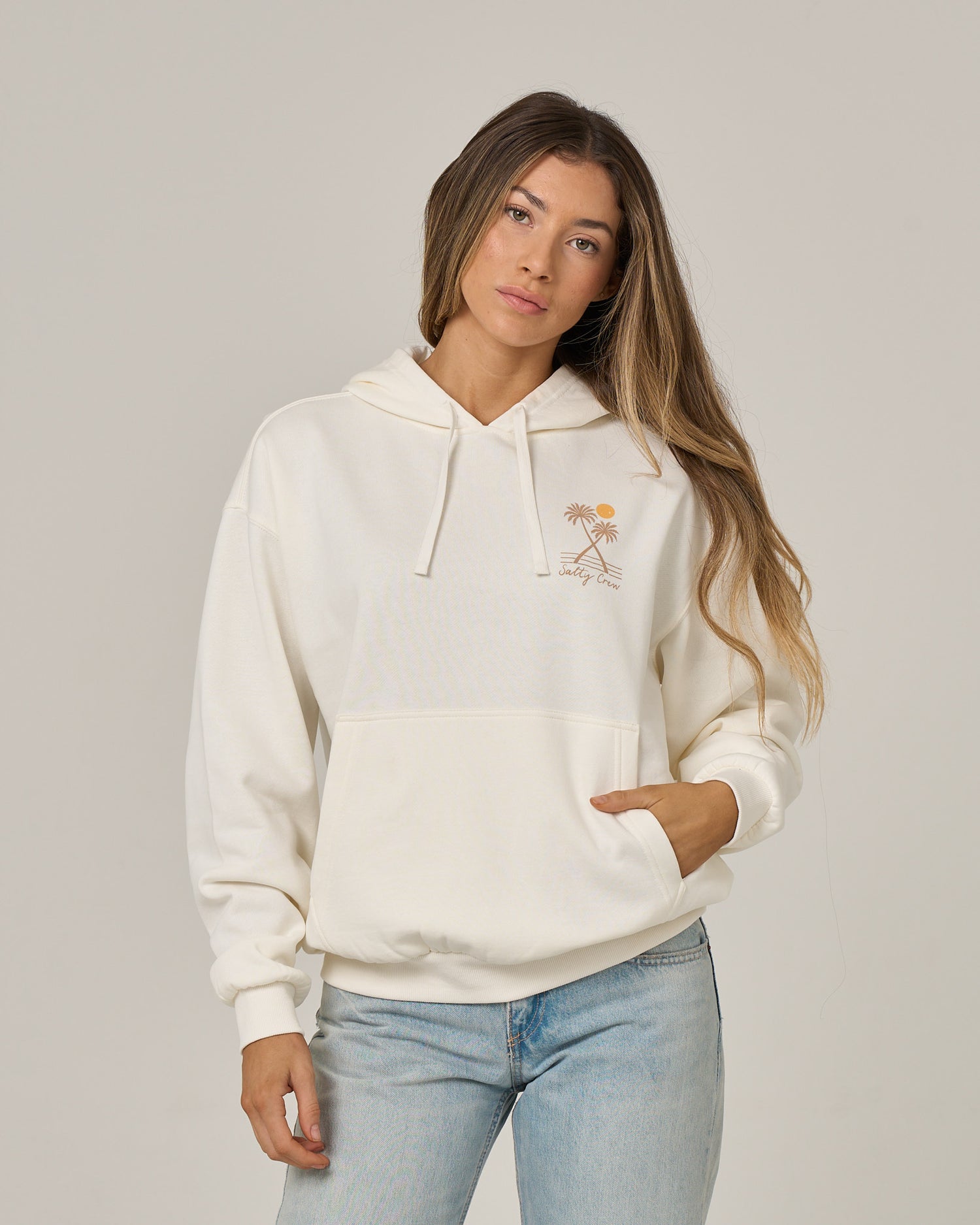 A woman with long hair wears the Salty Crew Moon Wake Hoodie in Off White, featuring a small orange palm tree graphic, paired with light blue jeans. She stands against a plain light background with one hand casually resting in the kangaroo pocket of her hoodie.
