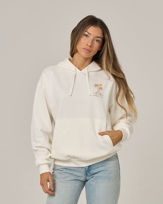 A woman with long hair wears the Salty Crew Moon Wake Hoodie in Off White, featuring a small orange palm tree graphic, paired with light blue jeans. She stands against a plain light background with one hand casually resting in the kangaroo pocket of her hoodie.