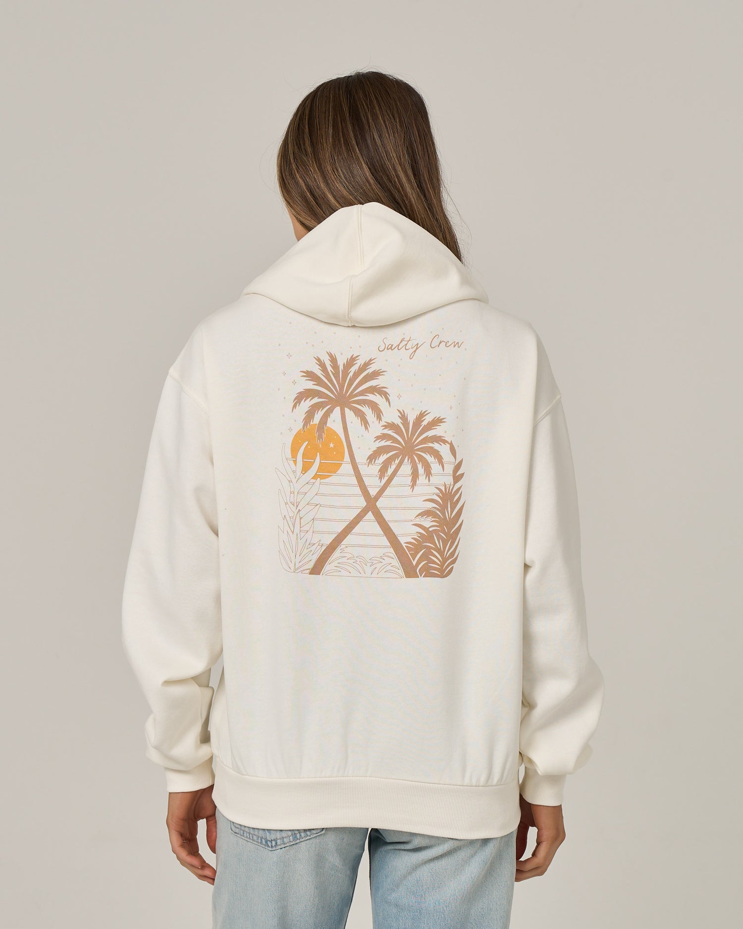 A person wearing the Salty Crew Moon Wake Hoodie in Off White, featuring an oversized fit with a graphic of palm trees and a sunset on the back. The text “Salty Crew” appears above the artwork. The fleece hoodie includes a classic kangaroo pocket, and the person is facing away, also dressed in light blue jeans.