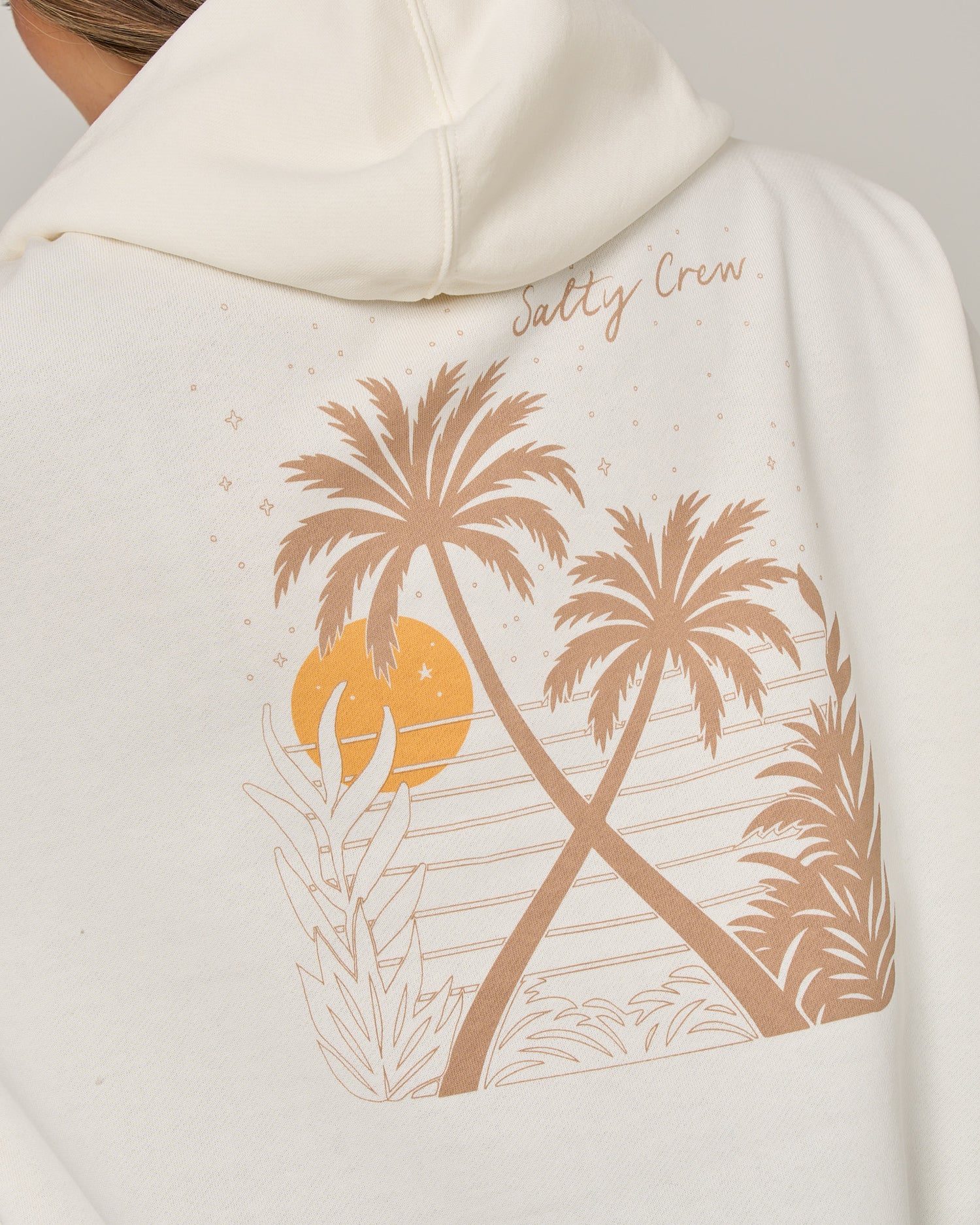 The Salty Crew Moon Wake Hoodie in Off White is an oversized fleece hoodie featuring a graphic of two palm trees, a setting sun, stars, and plants. It includes a cozy kangaroo pocket and has “Salty Crew” printed above the design on the back.