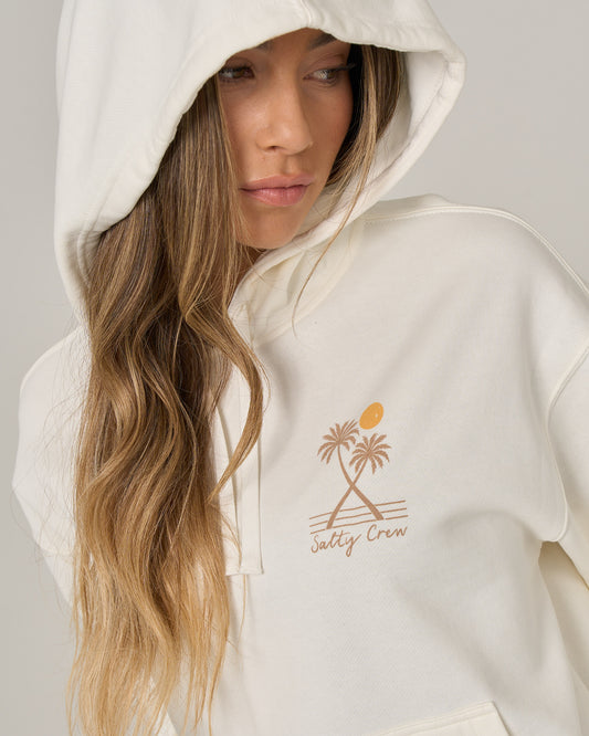 A person wearing the Salty Crew Moon Wake Hoodie in Off White, featuring a palm tree graphic, looks to the side. The hood is pulled up over part of their head, long hair drapes over their shoulder, and their hands rest casually in the kangaroo pocket.