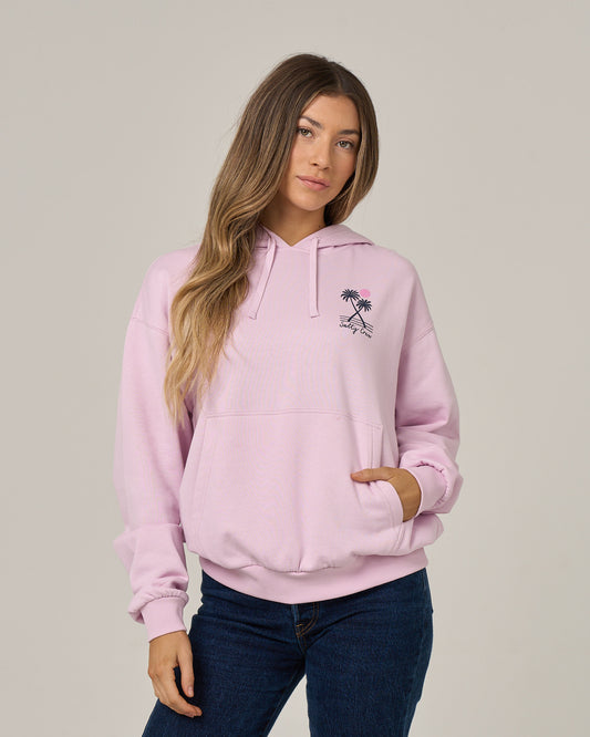 A woman wears the Salty Crew Moon Wake Hoodie in Pale Orchid—a light pink cotton fleece featuring a small palm tree graphic on the left chest and a cozy kangaroo pocket—which she pairs with blue jeans while standing against a plain light gray background.