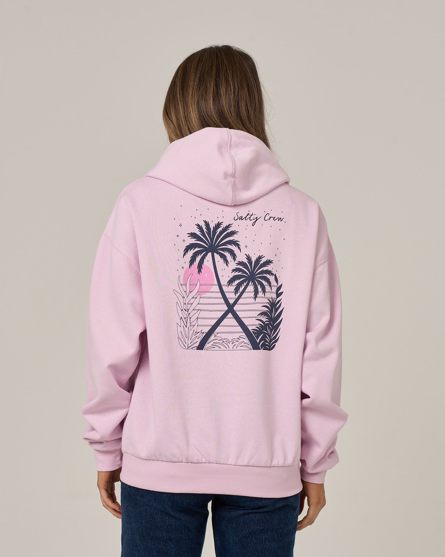 A person is seen from behind wearing the Salty Crew Moon Wake Hoodie in Pale Orchid, an oversized light pink cotton fleece sweatshirt featuring a tropical beach graphic with palm trees and waves, plus “Salty Crew” text on the back. The cozy hoodie includes a kangaroo pocket.