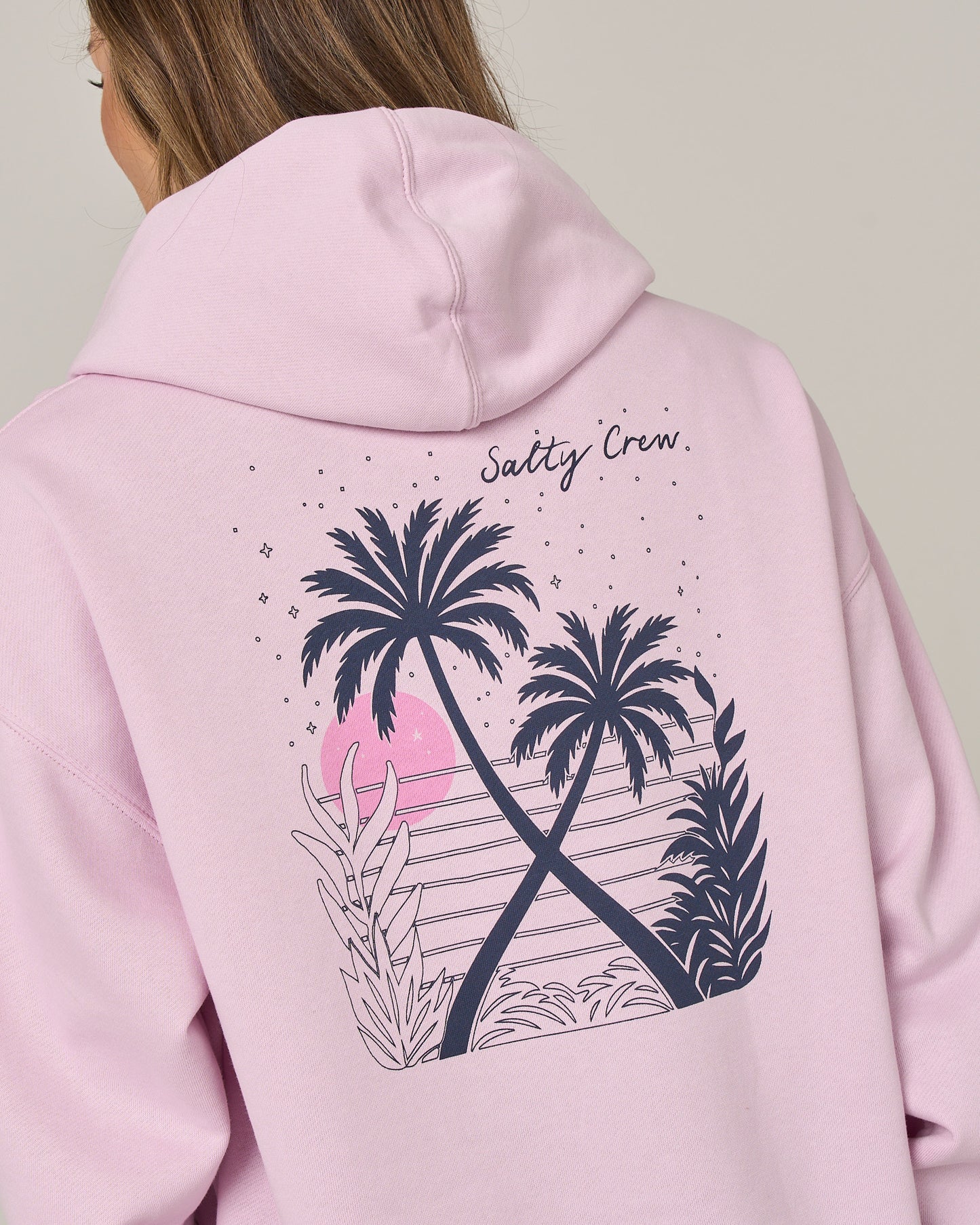 A person wearing the Salty Crew Moon Wake Hoodie in Pale Orchid—a cozy, oversized cotton fleece hoodie featuring a graphic of two palm trees, tropical plants, and a pink sun on the back, with “Salty Crew” text and a comfortable kangaroo pocket in front.