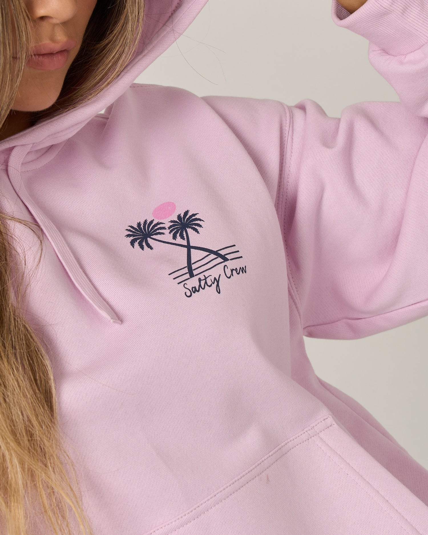 A person wearing the Salty Crew Moon Wake Hoodie in Pale Orchid—a soft cotton fleece, oversized style with a palm tree and sunset graphic and “Salty Crew” text on the chest—shows only part of their face and upper body. The hoodie features a classic kangaroo pocket.