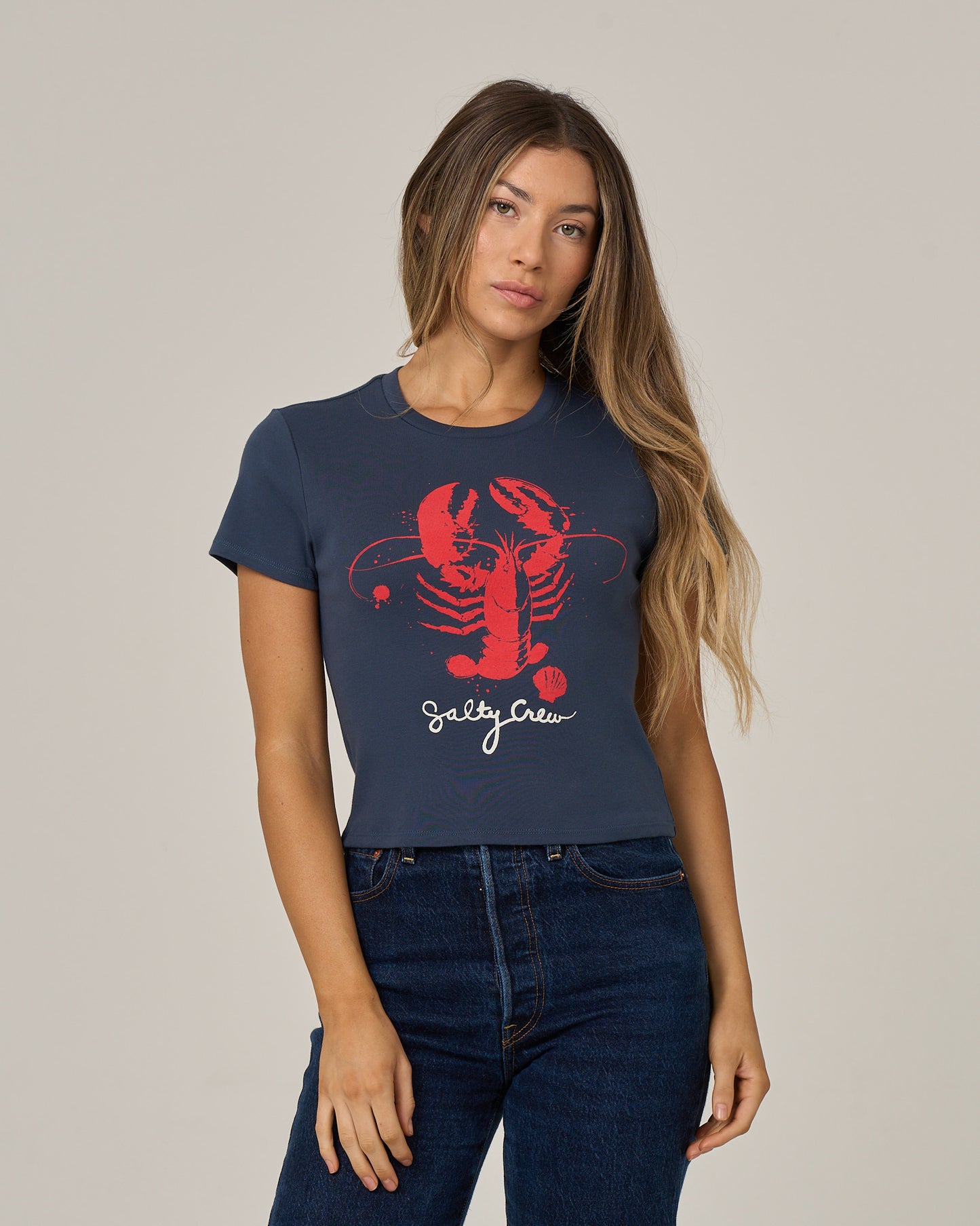 A woman with long hair wears the Salty Crew Captains Special Baby Tee in True Navy, a fitted short sleeve cotton shirt featuring a large red lobster graphic and bold Salty Crew lettering, paired with blue jeans and standing against a plain background.