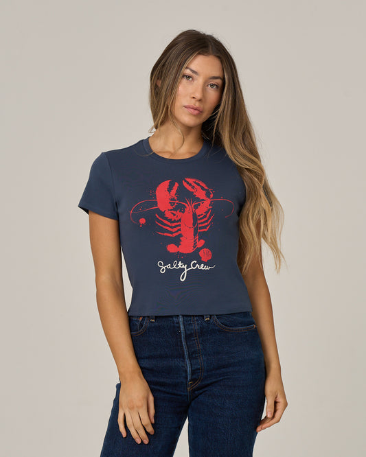 A woman with long hair wears the Salty Crew Captains Special Baby Tee in True Navy, a fitted short sleeve cotton shirt featuring a large red lobster graphic and bold Salty Crew lettering, paired with blue jeans and standing against a plain background.