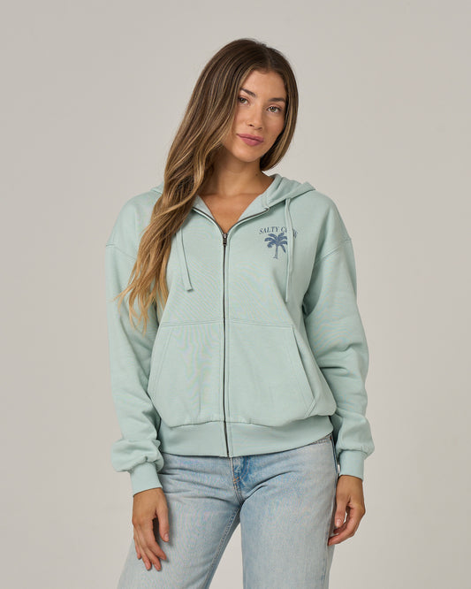 A woman with long brown hair models the Salty Crew Pretty Palm Hooded Zip in Cloud Blue—a relaxed fit, soft cotton-polyester fleece hoody with a zip front and a small navy palm tree logo on the chest. She pairs it with light-wash blue jeans against a plain gray background.